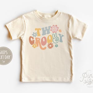 May include: A light beige short-sleeved t-shirt with the text "Two Groovy" in colorful retro lettering and a peace sign graphic.