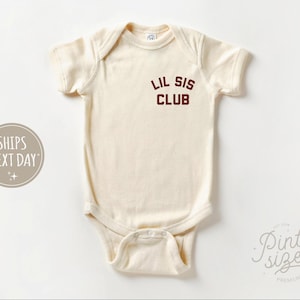 May include: Cream-colored baby onesie with short sleeves. The text "LIL SIS CLUB" is printed in maroon on the chest. The onesie has a snap closure at the bottom and a round neckline. The overall design is simple and minimalist.