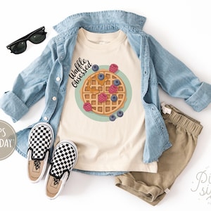 Peut inclure: Un t-shirt cr&egrave;me avec un dessin de gaufre, des myrtilles et des framboises, avec le texte "Waffle Obsessed". La tenue comprend une chemise en jean, un short kaki et des chaussures &agrave; enfiler &agrave; carreaux noirs et blancs. Des lunettes de soleil noires sont &eacute;galement incluses.