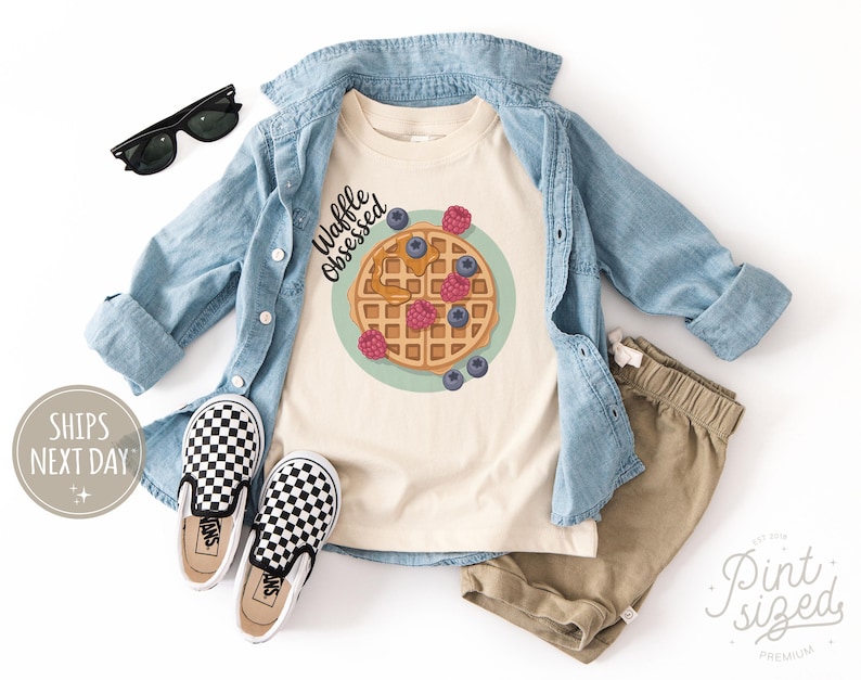 Peut inclure: Un t-shirt cr&egrave;me avec un dessin de gaufre, des myrtilles et des framboises, avec le texte "Waffle Obsessed". La tenue comprend une chemise en jean, un short kaki et des chaussures &agrave; enfiler &agrave; carreaux noirs et blancs. Des lunettes de soleil noires sont &eacute;galement incluses.