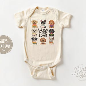 May include: Cream-colored baby bodysuit featuring a colorful graphic of nine different dog breeds. The phrase "ALL YOU NEED IS LOVE" is printed in black. The bodysuit has short sleeves and a snap closure.