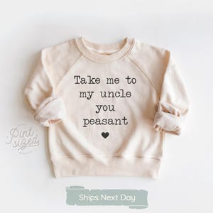 May include: Cream-colored sweatshirt with raglan sleeves. The front of the sweatshirt features the text "Take me to my uncle you peasant" in black, with a small black heart below the text. The sweatshirt is designed for children.