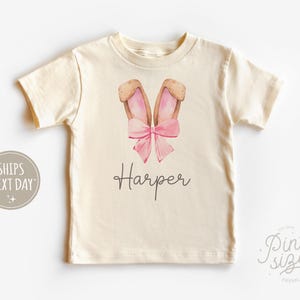 May include: A cream colored t-shirt with a pink bow and brown bunny ears. The name "Harper" is printed on the shirt.
