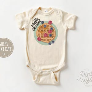 Peut inclure: Body de b&eacute;b&eacute; cr&egrave;me avec un motif de gaufre. La gaufre est garnie de sirop, de framboises et de myrtilles. Le texte "Waffle Obsessed" est au-dessus de la gaufre. Le body a des manches courtes et une fermeture &agrave; boutons-pression.