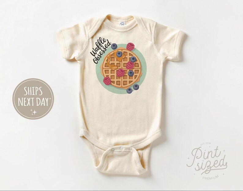 Peut inclure: Body de b&eacute;b&eacute; cr&egrave;me avec un motif de gaufre. La gaufre est garnie de sirop, de framboises et de myrtilles. Le texte "Waffle Obsessed" est au-dessus de la gaufre. Le body a des manches courtes et une fermeture &agrave; boutons-pression.