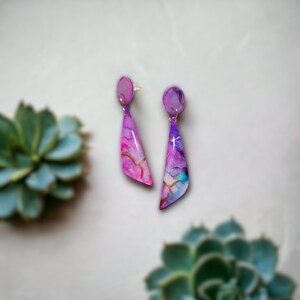 May include: A pair of dangle earrings with a purple, pink, and blue watercolor design and gold accents.