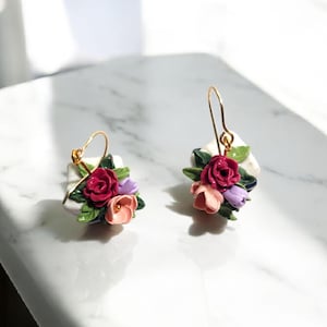 May include: A pair of floral earrings featuring a cluster of miniature roses in shades of red, pink, and purple, accented with green leaves. The flowers are set on a white square base with gold-colored ear wires.