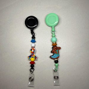 May include: Two retractable badge holders. One has a black reel with a colorful ribbon charm and beaded accents. The other has a mint green reel with a cowboy boot charm and beaded accents. Both have clear plastic clips.