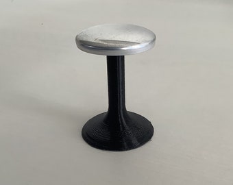 Può includere: Un piccolo oggetto autonomo caratterizzato da un disco superiore circolare in argento metallizzato, sostenuto da una base cilindrica verticale nera e testurizzata che si allarga sul fondo per formare una base circolare stabile.