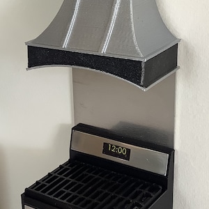May include: A miniature black stove with a silver control panel, a black grate top, and a digital display showing 12:00. Above the stove is a silver metallic backsplash and a silver range hood with a black textured band along the bottom edge.