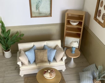 May include: A miniature living room scene featuring a cream-colored sofa with blue patterned and beige pillows, a round wooden coffee table with a bowl of seashells, a tall wooden shelving unit, a small side table with a lamp, and wall art.