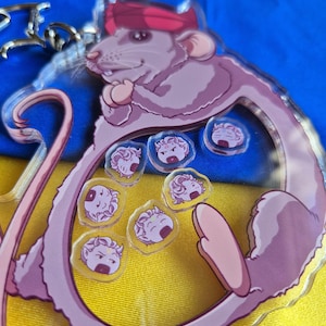 Puede incluir: Un llavero acrílico transparente con una rata de dibujos animados con una bandana roja. La rata es de color púrpura claro con una larga cola y está rodeada de pequeños amuletos circulares con varias expresiones faciales. El fondo es amarillo y azul.