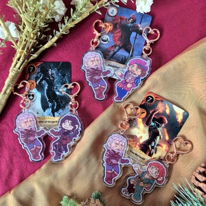 Peut inclure: Quatre porte-clés en acrylique représentant des personnages de dessins animés d'un jeu fantastique. Chaque porte-clés a un fermoir en forme de cœur et une illustration de personnage. Les porte-clés sont associés à des cartes de jeu. Le fond est un tissu bordeaux.