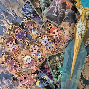 May include: A selection of character keychains and trading cards with fantasy artwork. The keychains have gold-coloured metal details and show cartoon-style characters. The trading cards display detailed illustrations of fantasy figures. The background is a patterned fabric.
