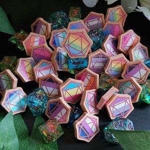 May include: A collection of polyhedral dice and wooden tokens with colorful geometric designs. The dice are clear with glitter and the tokens feature various pride flag patterns. The items are arranged on a dark surface with greenery and white flowers.