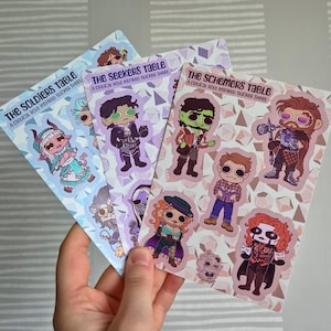 Peut inclure: Une main tient trois planches d'autocollants avec des personnages de dessins animés. Chaque planche a une palette de couleurs différente et le texte "THE SOLDIERS TABLE", "THE SEEKERS TABLE" et "THE SCHEMERS TABLE". Les autocollants sont inspirés de Critical Role.