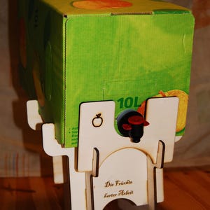 Puede incluir: Una caja de jugo verde de 10L con ilustraciones de manzanas, colocada en un dispensador de madera blanca. El dispensador tiene un grifo negro y rojo y el texto "Die Früchte harter Arbeit".