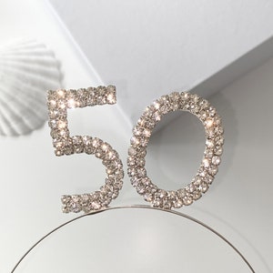 May include: A silver headband with the number 50 made of rhinestones. The headband is on a white surface.