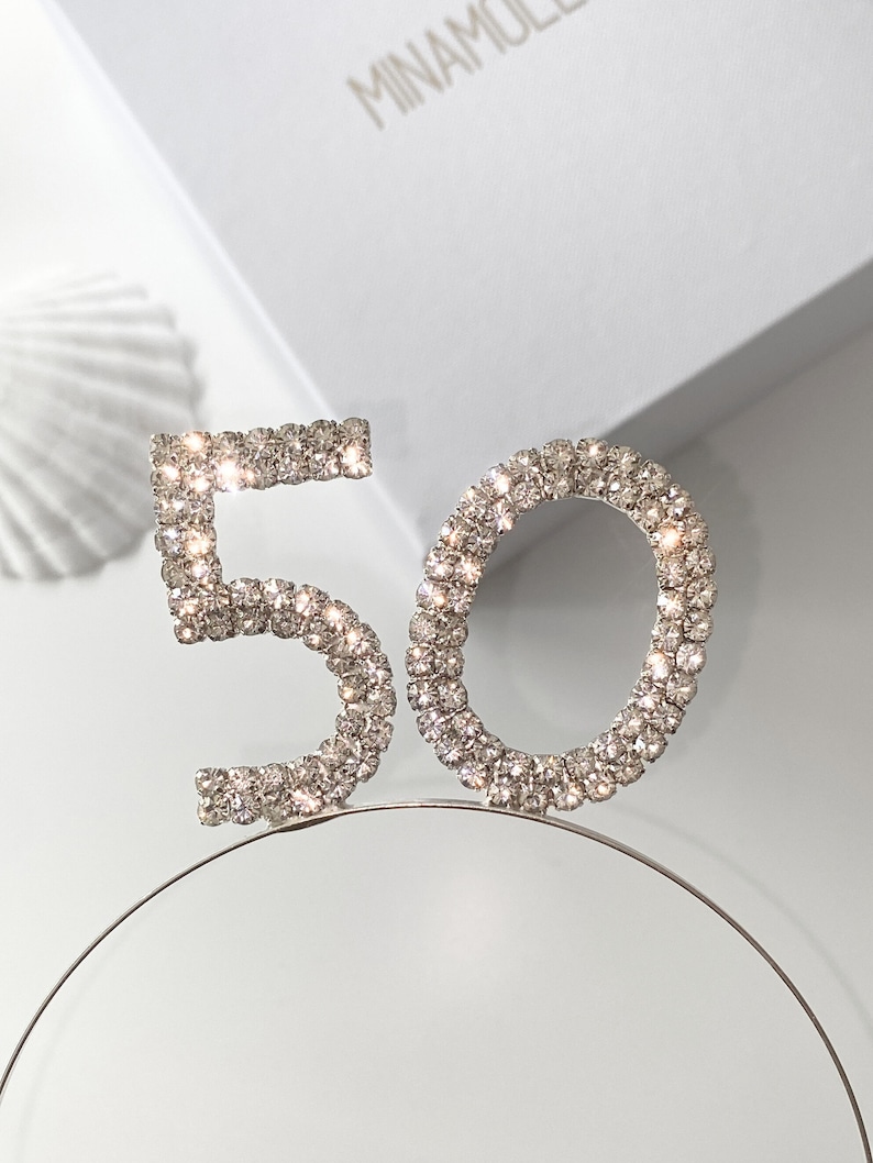 May include: A silver headband with the number 50 made of rhinestones. The headband is on a white surface.