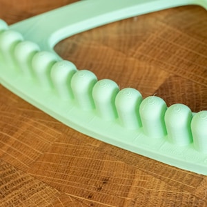 Op de afbeelding: Een lichtgroen plastic gereedschap met een rij ronde bulten langs de rand. Het gereedschap is driehoekig met een afgeronde rand.