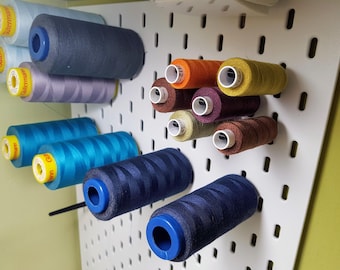May include: A white pegboard with various colors of thread spools organized by color. The spools are blue, teal, gray, orange, yellow, brown, and red.