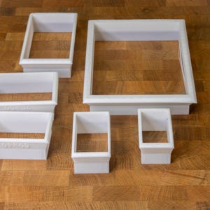 May include: Five white plastic cookie cutters of different sizes. The cutters are shaped like squares with rounded corners. The largest cutter is 10 inches wide and the smallest cutter is 3 inches wide.