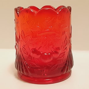 May include: A red glass toothpick holder with a cherry and leaf design. The holder has a scalloped top and a ribbed bottom.
