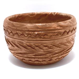 May include: A handcrafted, round, brown ceramic bowl with a textured surface. The bowl features carved patterns, including a zigzag design and a leaf-like texture. The interior is smooth, and the bowl is set against a white background.