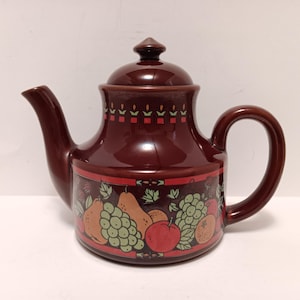May include: A maroon ceramic teapot with a lid and handle. The teapot features a fruit design with apples, pears, and grapes. The design is framed by a red border. The teapot is suitable for serving tea.