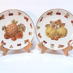 May include: Two decorative plates with fruit designs. One plate features a pineapple and strawberries, the other has pears and hazelnuts. Both plates have a gold rim and are displayed on wooden stands.