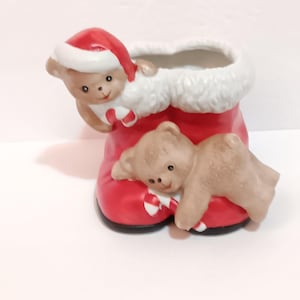 May include: A red ceramic Christmas boot with a white fluffy trim, decorated with two teddy bears. One bear wears a Santa hat and holds a candy cane. The other bear is lying on the boot. Festive holiday decor.