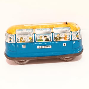 May include: Vintage toy bus in blue and yellow. The bus has windows with passengers inside. The bus has the numbers 1 and 2 on the side, and the text "GE 300". The bus has two axles with wheels.