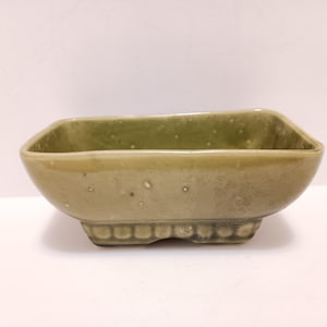 May include: A small, olive-green ceramic bowl with a rectangular shape and rounded corners. The bowl has a slightly textured surface with small, irregular spots. The base features a decorative, repeating pattern. The bowl is likely used for decorative purposes.
