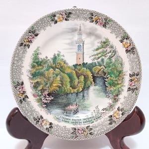 May include: Decorative plate featuring a scenic landscape with a river, trees, and a tall tower. The plate has a floral border and text that reads "Stephen Foster Memorial".