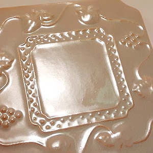 May include: A square ceramic tile with a raised design of a frame with grapes and leaves. The tile is a light brown color with a glossy finish.