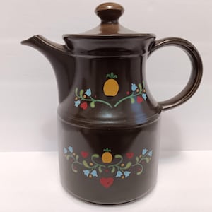 May include: A dark brown ceramic coffee pot with a lid and handle. The pot is decorated with a floral design featuring yellow pineapples, red hearts, and blue flowers. The spout is on the left side.