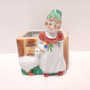 May include: A ceramic planter featuring a young girl in a red and white dress, holding flowers, and standing next to a white goose. The planter has a brown, wood-like texture and a green base.
