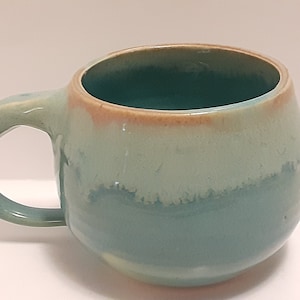 May include: A handmade ceramic mug with a rounded shape and a curved handle. The mug features a gradient of colors, including teal, light blue, and a touch of brown at the rim. This unique mug is perfect for enjoying your favorite hot beverages.