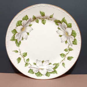 May include: A white ceramic plate with a gold rim, adorned with a floral design. The plate features three large white flowers with yellow centers and green leaves, arranged around the edge.