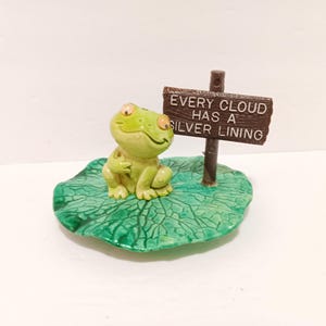 May include: A ceramic frog figurine with a sign that reads "Every cloud has a silver lining." The frog is light green and sitting on a green lily pad. The sign is brown with white lettering. A whimsical decorative item.