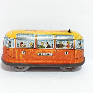 May include: Vintage toy bus in orange and yellow with passengers visible through the windows. The bus has two axles and the text "GE 300" is printed on the side. Numbers 1 and 2 are also visible on the bus.