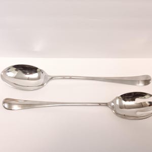 May include: A pair of shiny, silver serving utensils. One is a large spoon, and the other is a serving fork with three tines. Both have long, slender handles and are set against a white background.