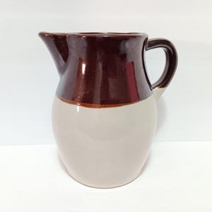 May include: A two-tone ceramic pitcher with a handle and spout. The top half is a dark brown, while the bottom half is a light beige. A thin brown band separates the two colors. The pitcher is likely used for serving beverages.