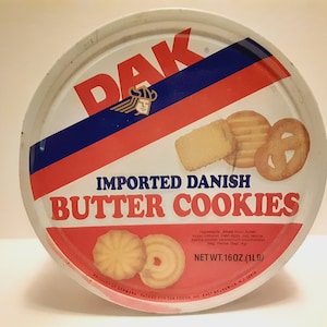 May include: A red, white, and blue tin can with the words "DAK Imported Danish Butter Cookies" printed on it. The can has a picture of a viking helmet and wings on it. The can also has a picture of cookies on it. The can is 16 ounces (1 pound).