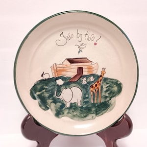 May include: A ceramic plate with a green rim and a hand-painted scene. The plate features an ark, animals, and the text "Two by two!". The animals include an elephant, giraffe, rabbit, and sheep.