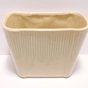 May include: Cream-colored rectangular ceramic planter with vertical ribbed texture on the sides. The planter has a slightly flared shape and a smooth, rounded rim. The interior is a lighter shade of cream.