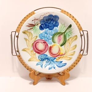 May include: A round, white ceramic serving platter with a hand-painted fruit design. The platter features a woven rattan border and metal handles. The fruit includes grapes, peaches, and a fig. The platter is displayed on a wooden stand.
