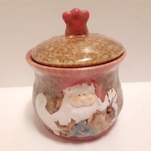 May include: A ceramic jar with a lid featuring a Santa Claus design. The jar is decorated with a brown and pink glaze and has a red knob on the lid.