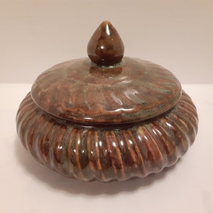 May include: A ceramic lidded jar with a brown and green glaze. The jar has a rounded body with a ribbed design and a domed lid with a pointed knob.