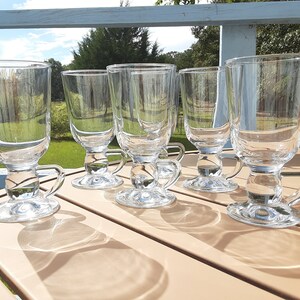 May include: Six clear glass mugs with handles, each with a rounded base and a flared top. The mugs are arranged in a row on a light brown wooden table.